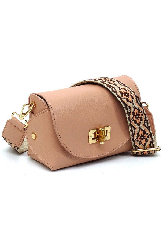 The Bamboo Twist Lock Flap Crossbody Bag is a small peach-colored bag made from faux vegan leather. It features a chic bamboo twist lock for added elegance, a gold clasp, and an adjustable patterned shoulder strap with black and brown geometric designs.