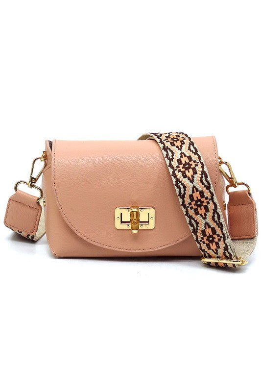 The Bamboo Twist Lock Flap Crossbody Bag is a small peach-colored bag made from faux vegan leather. It features a chic bamboo twist lock for added elegance, a gold clasp, and an adjustable patterned shoulder strap with black and brown geometric designs.