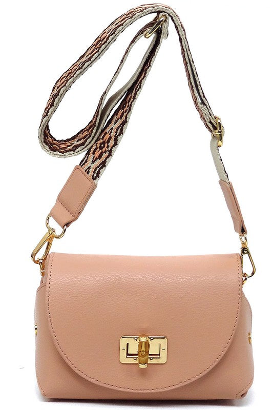 The Bamboo Twist Lock Flap Crossbody Bag is a small peach-colored bag made from faux vegan leather. It features a chic bamboo twist lock for added elegance, a gold clasp, and an adjustable patterned shoulder strap with black and brown geometric designs.