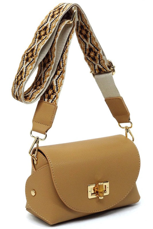 The Bamboo Twist Lock Flap Crossbody Bag is a small peach-colored bag made from faux vegan leather. It features a chic bamboo twist lock for added elegance, a gold clasp, and an adjustable patterned shoulder strap with black and brown geometric designs.