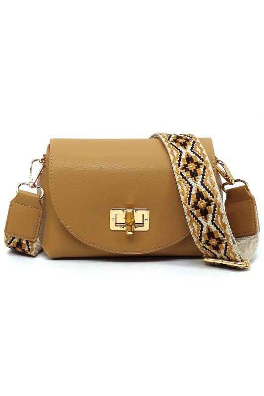 The Bamboo Twist Lock Flap Crossbody Bag is a small peach-colored bag made from faux vegan leather. It features a chic bamboo twist lock for added elegance, a gold clasp, and an adjustable patterned shoulder strap with black and brown geometric designs.