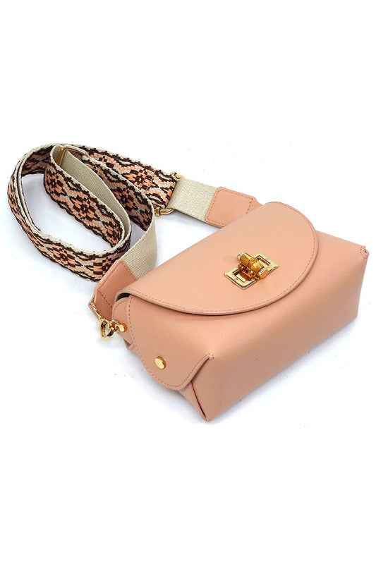 The Bamboo Twist Lock Flap Crossbody Bag is a small peach-colored bag made from faux vegan leather. It features a chic bamboo twist lock for added elegance, a gold clasp, and an adjustable patterned shoulder strap with black and brown geometric designs.