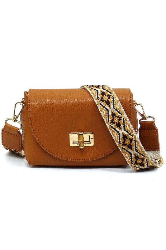 The Bamboo Twist Lock Flap Crossbody Bag is a small peach-colored bag made from faux vegan leather. It features a chic bamboo twist lock for added elegance, a gold clasp, and an adjustable patterned shoulder strap with black and brown geometric designs.