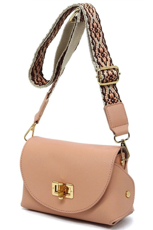 The Bamboo Twist Lock Flap Crossbody Bag is a small peach-colored bag made from faux vegan leather. It features a chic bamboo twist lock for added elegance, a gold clasp, and an adjustable patterned shoulder strap with black and brown geometric designs.