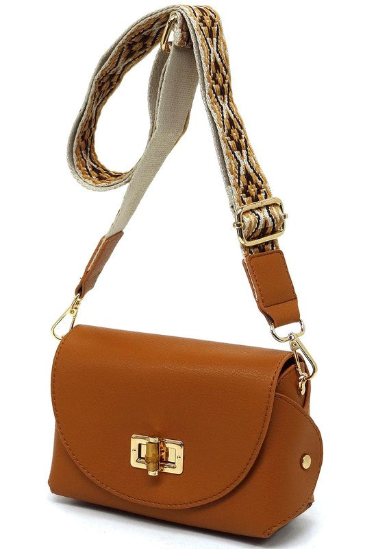 The Bamboo Twist Lock Flap Crossbody Bag is a small peach-colored bag made from faux vegan leather. It features a chic bamboo twist lock for added elegance, a gold clasp, and an adjustable patterned shoulder strap with black and brown geometric designs.