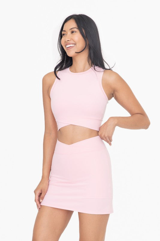 A woman is wearing a light pink Venice Crossover Active Top, which is sleeveless and has a high neck design. She pairs it with a matching skirt, all set against a plain white background. The ensemble, crafted from four-way stretch Venice fabric, effortlessly blends style with comfort.