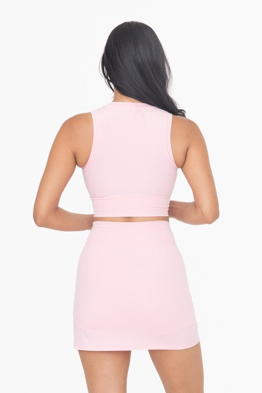 A woman is wearing a light pink Venice Crossover Active Top, which is sleeveless and has a high neck design. She pairs it with a matching skirt, all set against a plain white background. The ensemble, crafted from four-way stretch Venice fabric, effortlessly blends style with comfort.