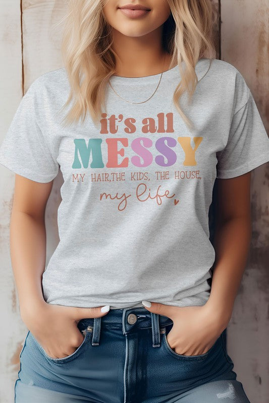 A person wearing a white "Mama Mom Mother's Day Graphic Tee" featuring the text "It's all messy my hair, the kids, the house, my life." The T-shirt is paired with blue jeans.