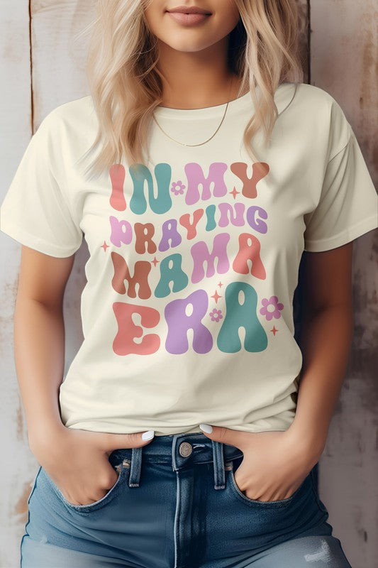 A woman in a Mama Mom Mother's Day Graphic Tee featuring the colorful text "IN MY PRAYING MAMA ERA," stands with her hands in her jeans pockets against a wooden background.