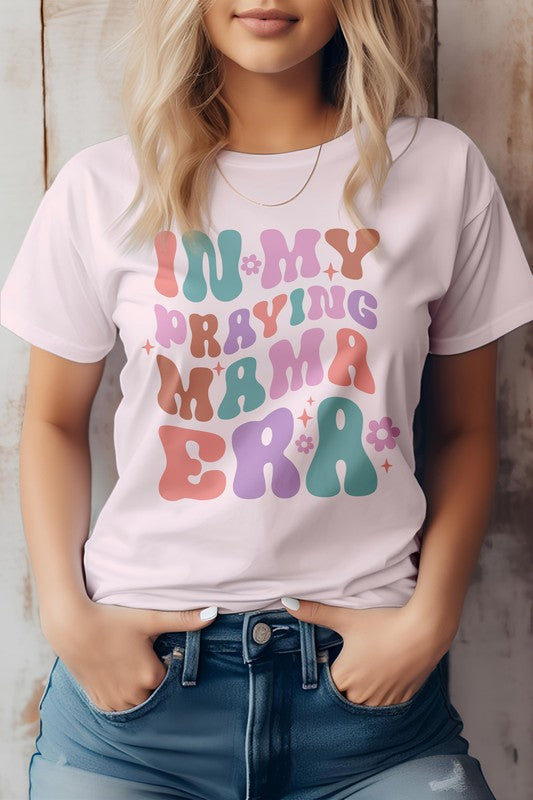 A woman in a Mama Mom Mother's Day Graphic Tee featuring the colorful text "IN MY PRAYING MAMA ERA," stands with her hands in her jeans pockets against a wooden background.