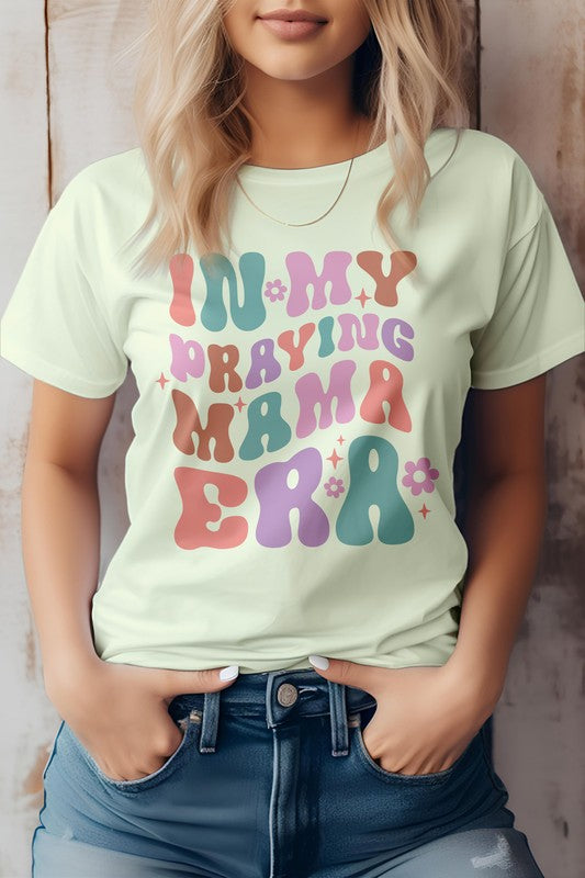 A woman in a Mama Mom Mother's Day Graphic Tee featuring the colorful text "IN MY PRAYING MAMA ERA," stands with her hands in her jeans pockets against a wooden background.