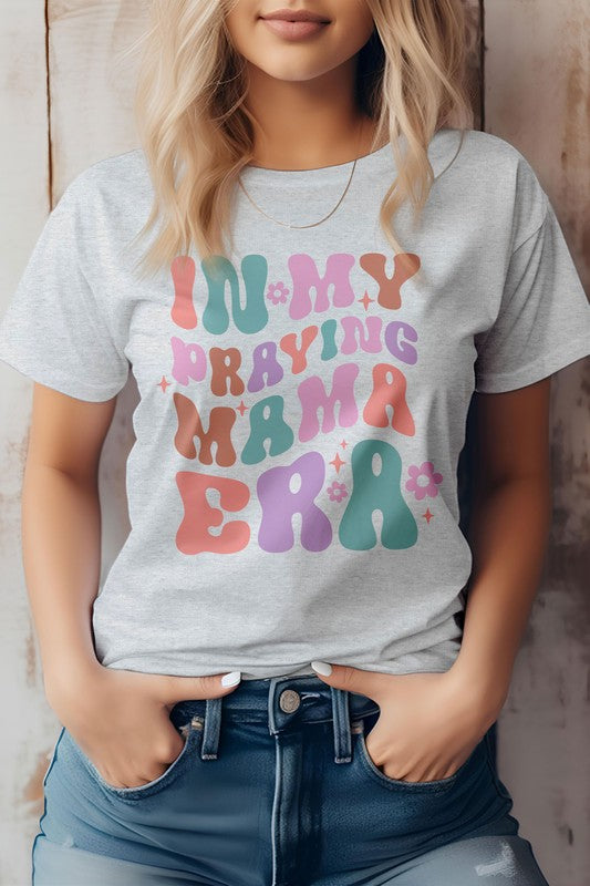 A woman in a Mama Mom Mother's Day Graphic Tee featuring the colorful text "IN MY PRAYING MAMA ERA," stands with her hands in her jeans pockets against a wooden background.