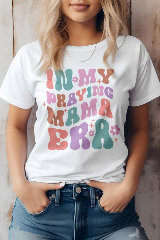 A woman in a Mama Mom Mother's Day Graphic Tee featuring the colorful text "IN MY PRAYING MAMA ERA," stands with her hands in her jeans pockets against a wooden background.