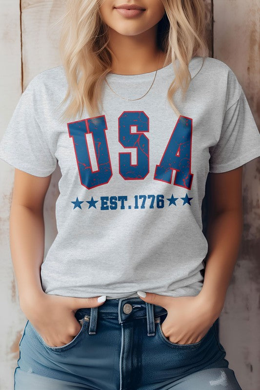 A woman wearing the "USA Retro America Est 1776 Graphic Tee," a light gray T-shirt featuring "USA EST. 1776" in bold blue and red letters, stands against a wooden background. Celebrating Independence Day, she has her hands in the pockets of her jeans, embracing patriotism with style and eco-friendly materials.