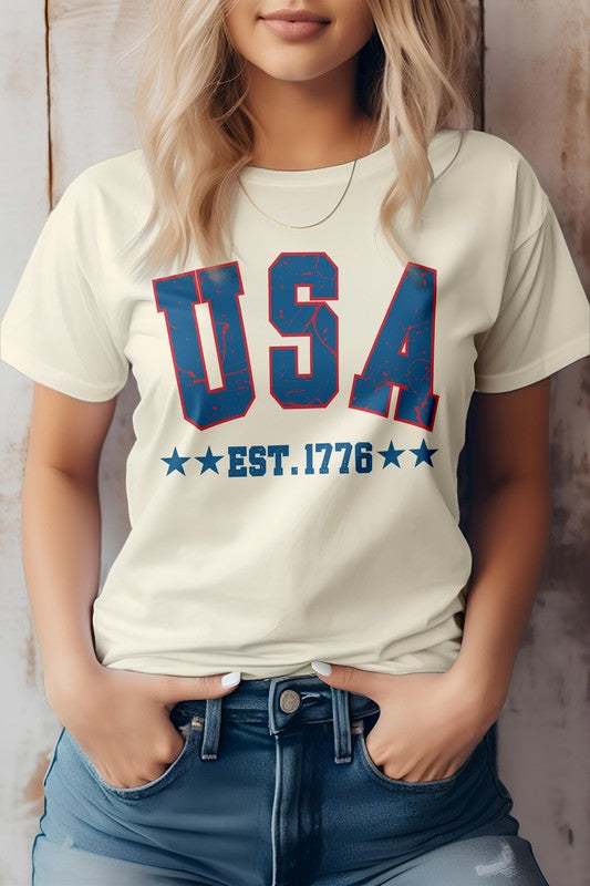 A woman wearing the "USA Retro America Est 1776 Graphic Tee," a light gray T-shirt featuring "USA EST. 1776" in bold blue and red letters, stands against a wooden background. Celebrating Independence Day, she has her hands in the pockets of her jeans, embracing patriotism with style and eco-friendly materials.