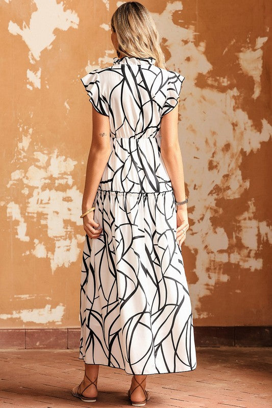 A woman is modeling the Ruffle Hem Loose Neck Elegant Ruffle Sleeve Dress, showcasing its white fabric adorned with a black abstract pattern, complete with ruffle details and a V-neckline, in front of an aged, textured wall. She accessorizes with a woven handbag and flat sandals.