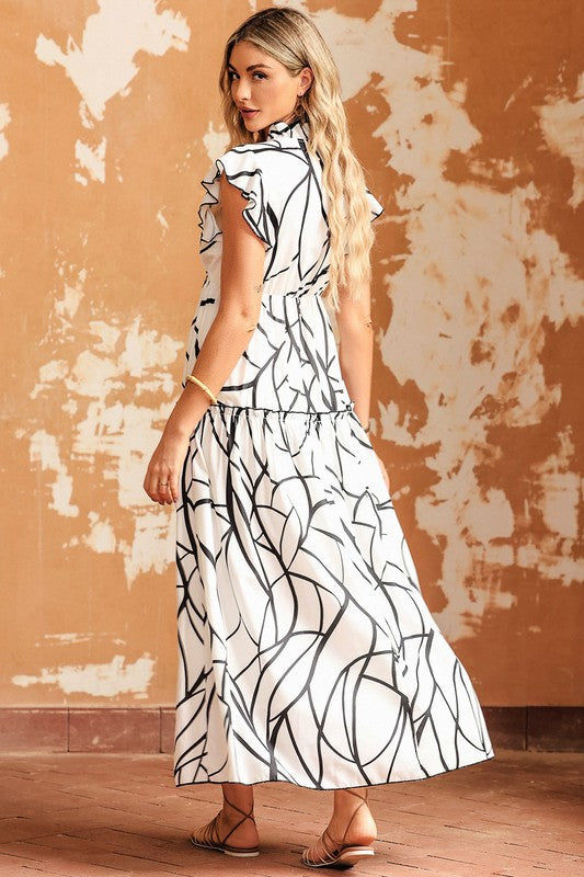 A woman is modeling the Ruffle Hem Loose Neck Elegant Ruffle Sleeve Dress, showcasing its white fabric adorned with a black abstract pattern, complete with ruffle details and a V-neckline, in front of an aged, textured wall. She accessorizes with a woven handbag and flat sandals.