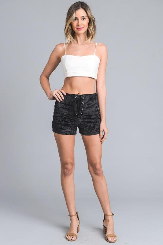 A person wearing black, stretch velvet Shorts with a lace-up front detail and beige open-toe heels. The upper body is partially visible.