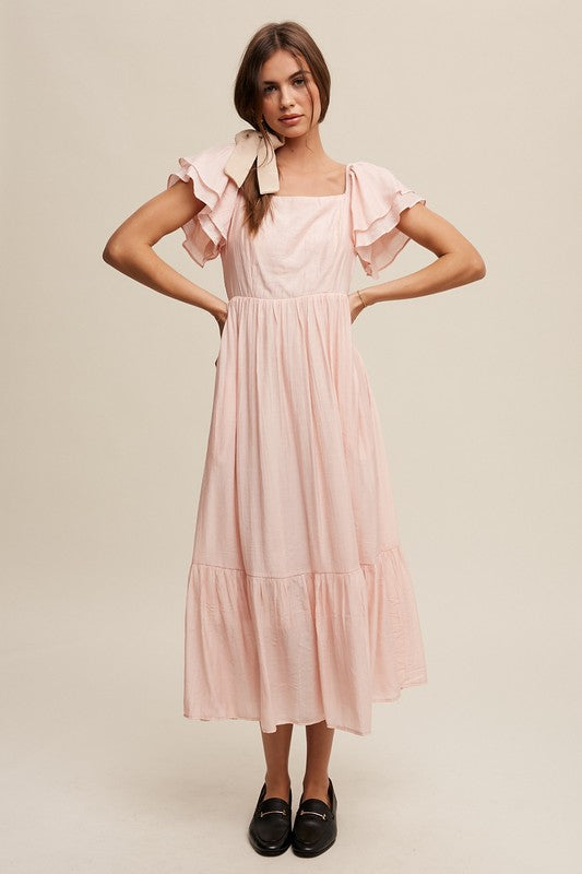 A woman wearing a Square Neck Ruffled Short Sleeve Maxi Dress in light pink, featuring crinkle textured fabric and a bow in her hair, stands gracefully against a neutral background.