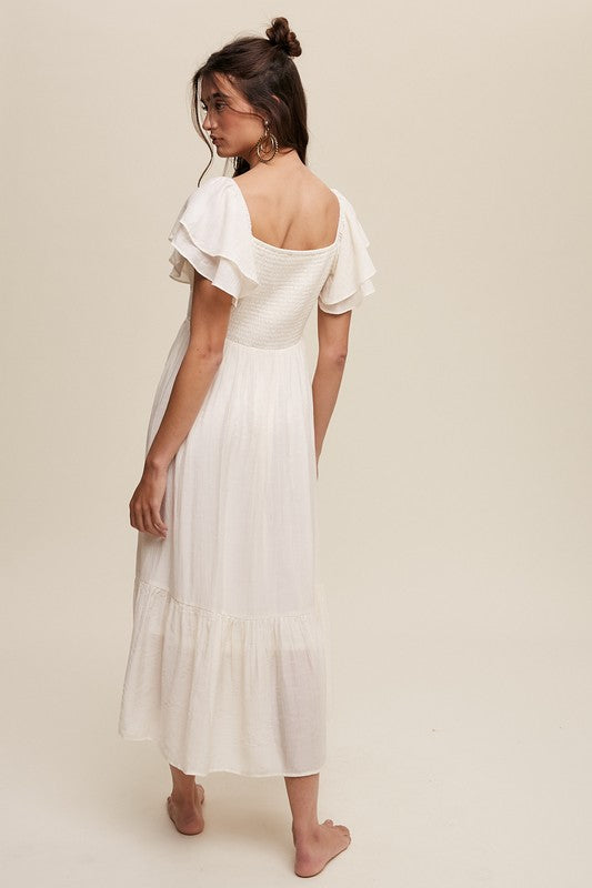 A woman wearing a Square Neck Ruffled Short Sleeve Maxi Dress in light pink, featuring crinkle textured fabric and a bow in her hair, stands gracefully against a neutral background.