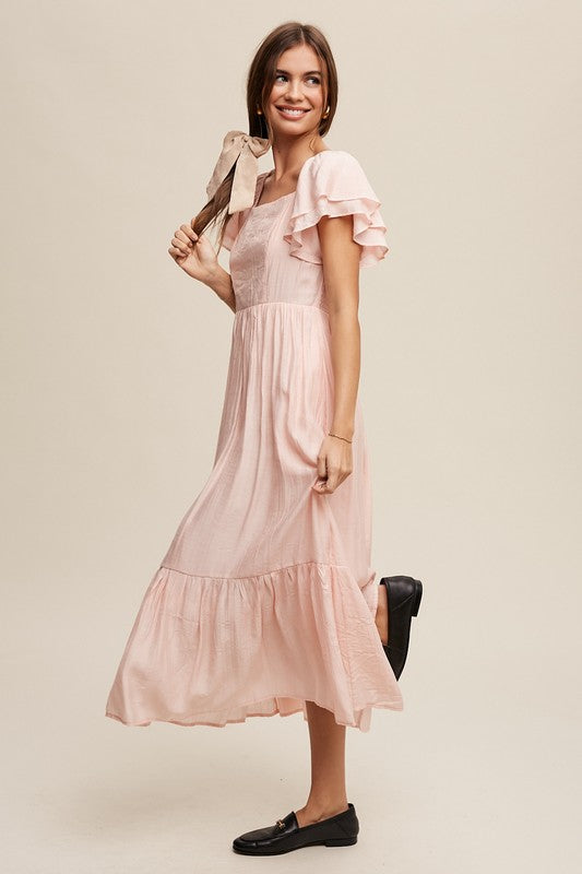 A woman wearing a Square Neck Ruffled Short Sleeve Maxi Dress in light pink, featuring crinkle textured fabric and a bow in her hair, stands gracefully against a neutral background.