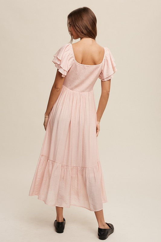 A woman wearing a Square Neck Ruffled Short Sleeve Maxi Dress in light pink, featuring crinkle textured fabric and a bow in her hair, stands gracefully against a neutral background.