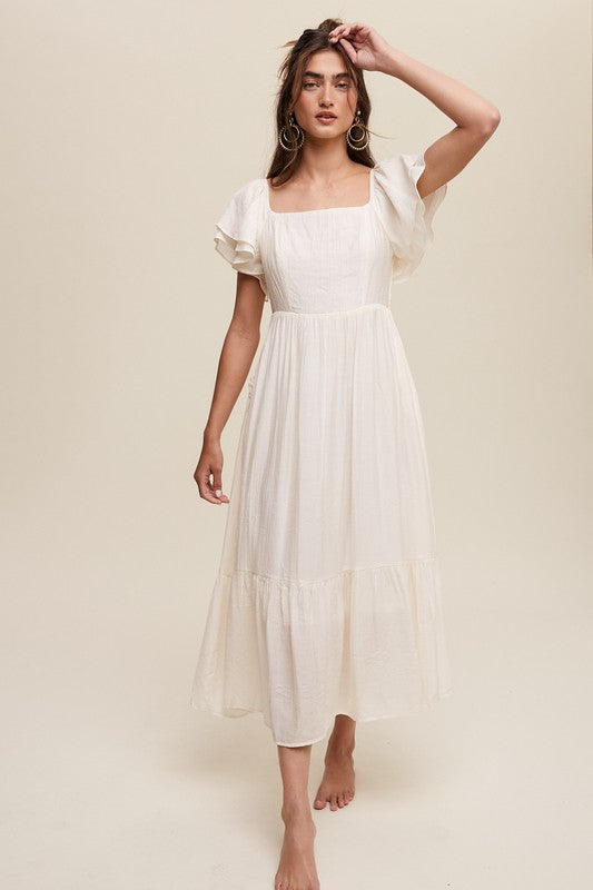 A woman wearing a Square Neck Ruffled Short Sleeve Maxi Dress in light pink, featuring crinkle textured fabric and a bow in her hair, stands gracefully against a neutral background.