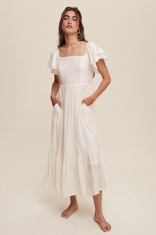 A woman wearing a Square Neck Ruffled Short Sleeve Maxi Dress in light pink, featuring crinkle textured fabric and a bow in her hair, stands gracefully against a neutral background.