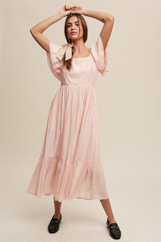 A woman wearing a Square Neck Ruffled Short Sleeve Maxi Dress in light pink, featuring crinkle textured fabric and a bow in her hair, stands gracefully against a neutral background.