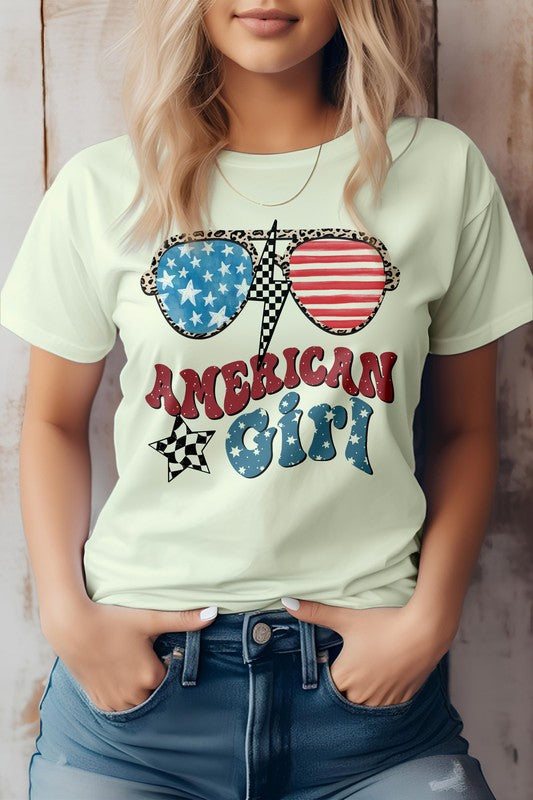 A woman with blonde hair is posing with her hands in her pockets, wearing an "American Girl, 4th of July Graphic Tee." She complements her look with sunglasses adorned in Patriotic USA Flag Glasses and the text "American Girl," making her outfit perfect for celebrating the 4th of July.