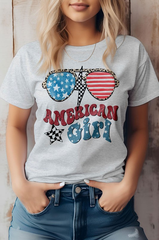 A woman with blonde hair is posing with her hands in her pockets, wearing an "American Girl, 4th of July Graphic Tee." She complements her look with sunglasses adorned in Patriotic USA Flag Glasses and the text "American Girl," making her outfit perfect for celebrating the 4th of July.