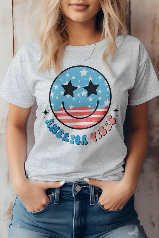 A person is wearing the America Vibes Retro, 4th of July Graphic Tee, showcasing a gray T-shirt with a retro American flag smiley face design featuring stars and stripes, along with the vibrant text "America Vibes." Ideal for celebrating the 4th of July, it is also made from eco-friendly materials.