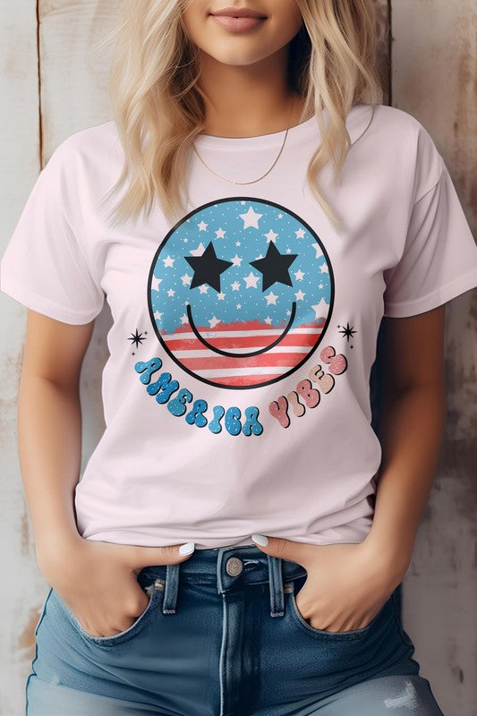 A person is wearing the America Vibes Retro, 4th of July Graphic Tee, showcasing a gray T-shirt with a retro American flag smiley face design featuring stars and stripes, along with the vibrant text "America Vibes." Ideal for celebrating the 4th of July, it is also made from eco-friendly materials.