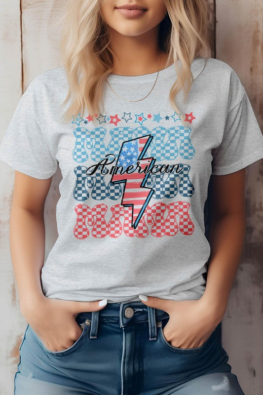 A person wearing the "American Mama Retro" graphic tee, featuring blue and red checkered letters with a lightning bolt design, tucked into jeans.