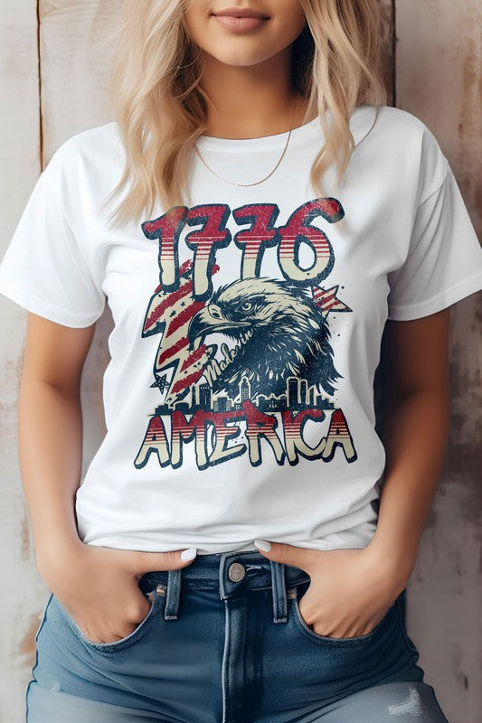 Against a wooden backdrop, an individual wears the 1776 America 4th of July Oversized Tee, crafted from eco-friendly materials. This shirt features "1776" and an eagle graphic with the word "America," perfectly capturing the patriotic spirit of Independence Day.