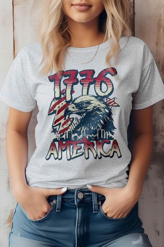 Against a wooden backdrop, an individual wears the 1776 America 4th of July Oversized Tee, crafted from eco-friendly materials. This shirt features "1776" and an eagle graphic with the word "America," perfectly capturing the patriotic spirit of Independence Day.