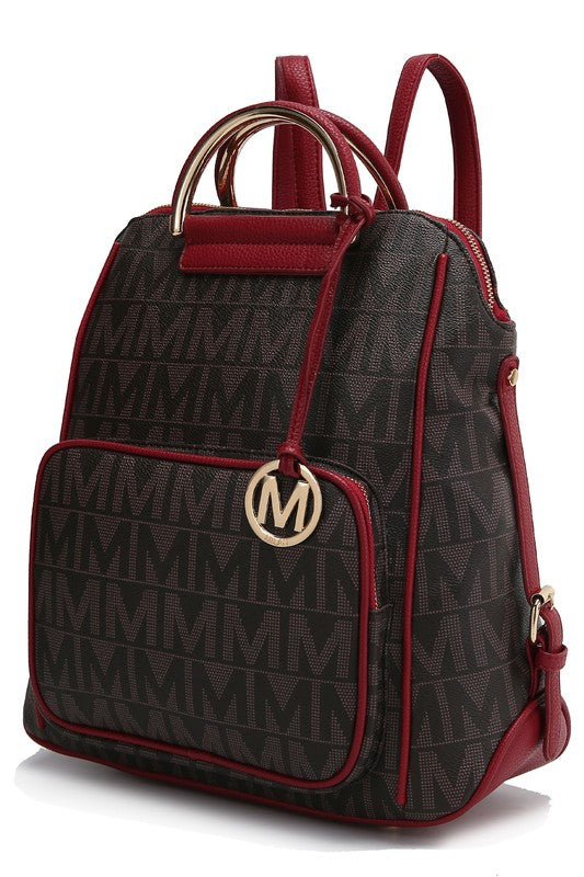 A person wearing a light-colored jacket and floral skirt is carrying the MKF Collection Cora Milan Backpack by Mia K, which features a blue pattern with metallic accents, an 'M' logo, and an adjustable shoulder strap.