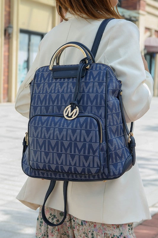 A person wearing a light-colored jacket and floral skirt is carrying the MKF Collection Cora Milan Backpack by Mia K, which features a blue pattern with metallic accents, an 'M' logo, and an adjustable shoulder strap.