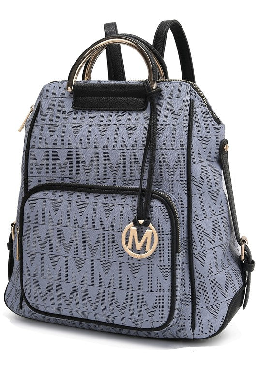 A person wearing a light-colored jacket and floral skirt is carrying the MKF Collection Cora Milan Backpack by Mia K, which features a blue pattern with metallic accents, an 'M' logo, and an adjustable shoulder strap.