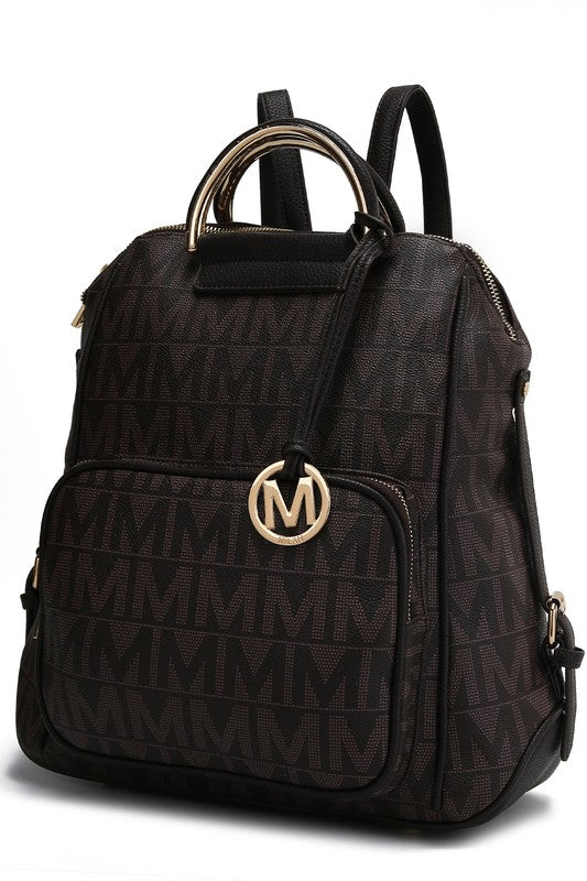 A person wearing a light-colored jacket and floral skirt is carrying the MKF Collection Cora Milan Backpack by Mia K, which features a blue pattern with metallic accents, an 'M' logo, and an adjustable shoulder strap.