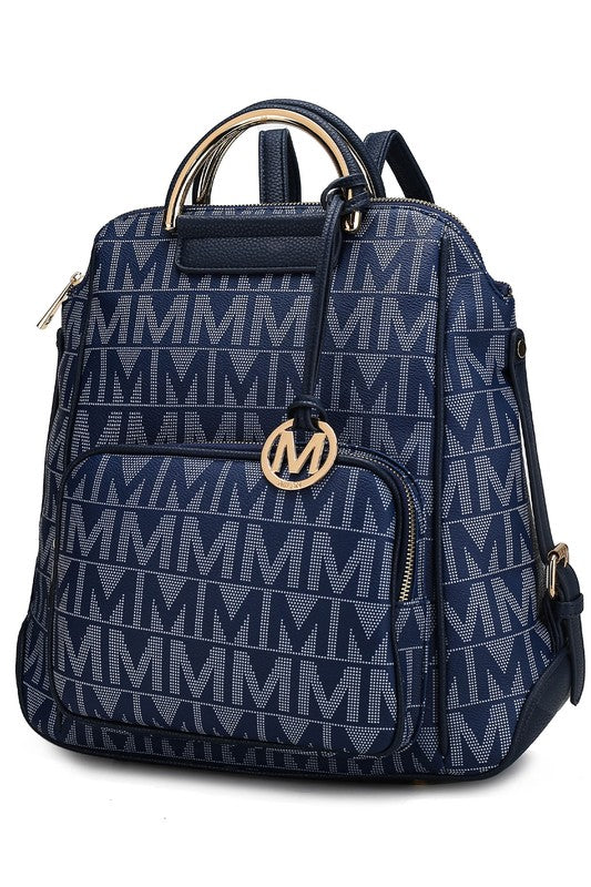 A person wearing a light-colored jacket and floral skirt is carrying the MKF Collection Cora Milan Backpack by Mia K, which features a blue pattern with metallic accents, an 'M' logo, and an adjustable shoulder strap.