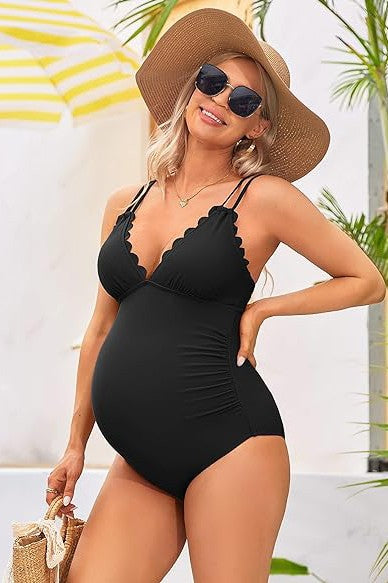 A pregnant woman in a Ruch Scallop V Neck One Piece Maternity Swimsuit, holding her belly, stands smiling in an outdoor setting with wooden and white walls in the background.