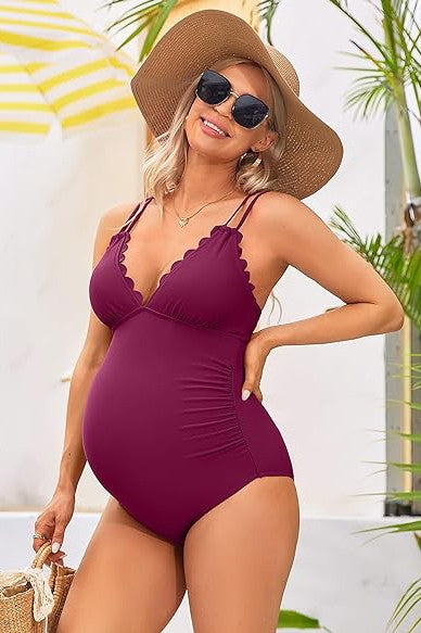 A pregnant woman in a Ruch Scallop V Neck One Piece Maternity Swimsuit, holding her belly, stands smiling in an outdoor setting with wooden and white walls in the background.