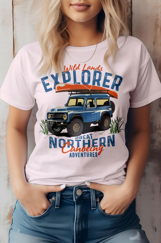 A person is wearing the Wild Lands Explorer, Adventure Graphic Tee, which showcases a vintage 4x4 design with a blue off-road vehicle and a canoe. The shirt features the text "Great Northern Canoeing Adventures" and is made from eco-friendly materials.