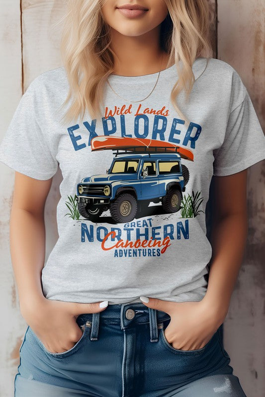 A person is wearing the Wild Lands Explorer, Adventure Graphic Tee, which showcases a vintage 4x4 design with a blue off-road vehicle and a canoe. The shirt features the text "Great Northern Canoeing Adventures" and is made from eco-friendly materials.