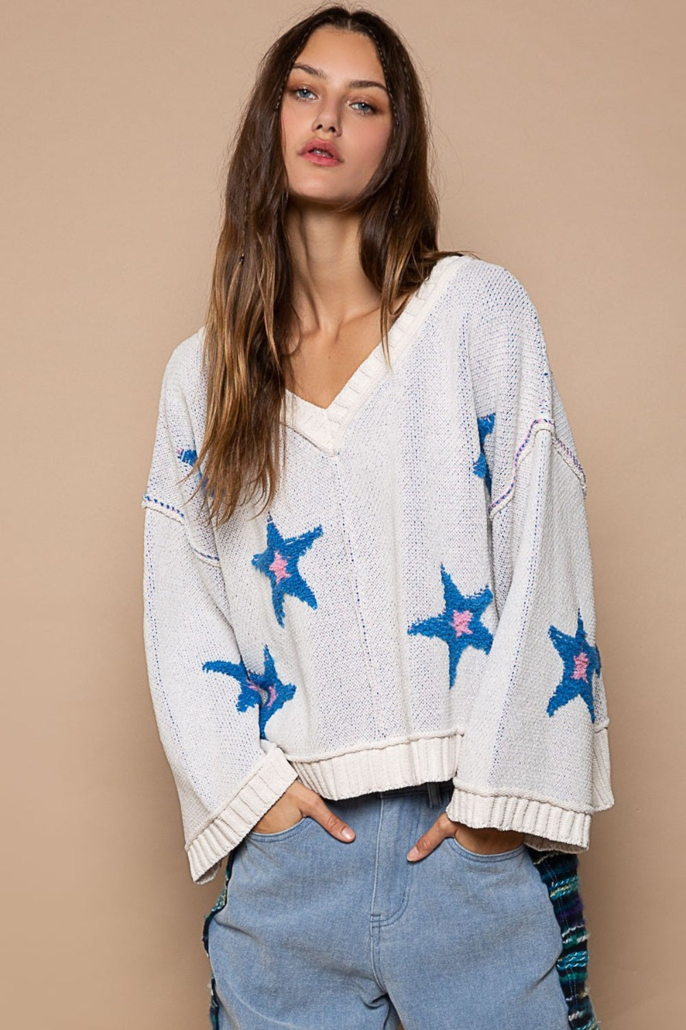 Woman in a POL Long Sleeve Star Patch Sweater with blue color-blocked star berber patches and light blue jeans standing against a beige background.