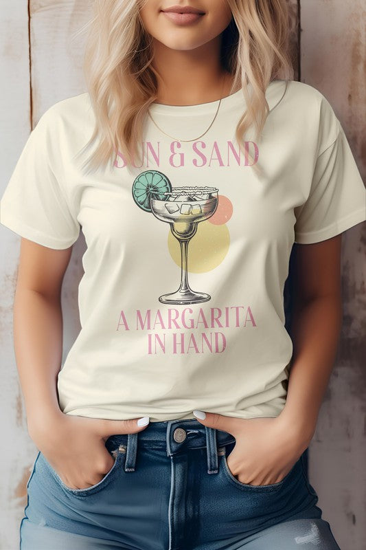 A woman wearing a Vintage Summer Beach Graphic Tee, featuring the phrase "Sun & Sand, A Margarita In Hand," crafted from eco-friendly materials, pairs it with denim shorts while standing against a wooden backdrop.