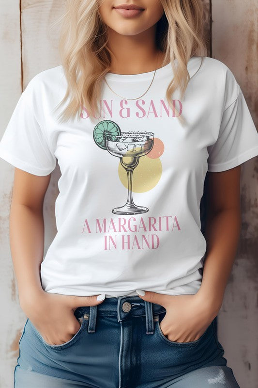 A woman wearing a Vintage Summer Beach Graphic Tee, featuring the phrase "Sun & Sand, A Margarita In Hand," crafted from eco-friendly materials, pairs it with denim shorts while standing against a wooden backdrop.