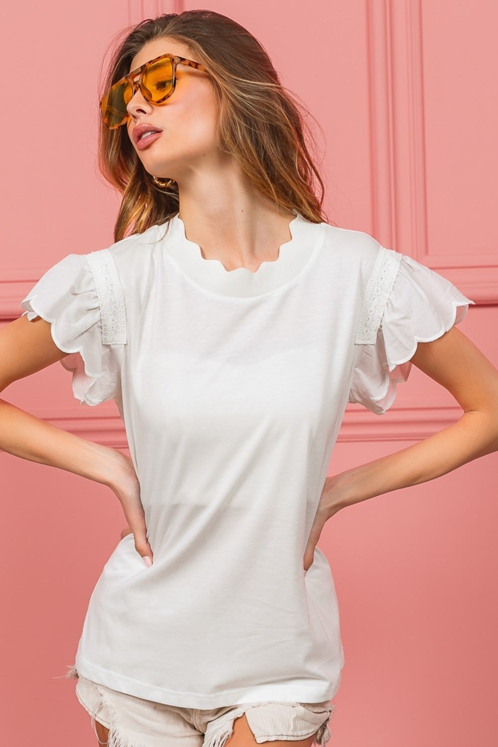 A woman with wavy hair dons the elegant BiBi Scallop Edged Lace Trim Top and large amber sunglasses while standing in front of a vibrant pink wall.