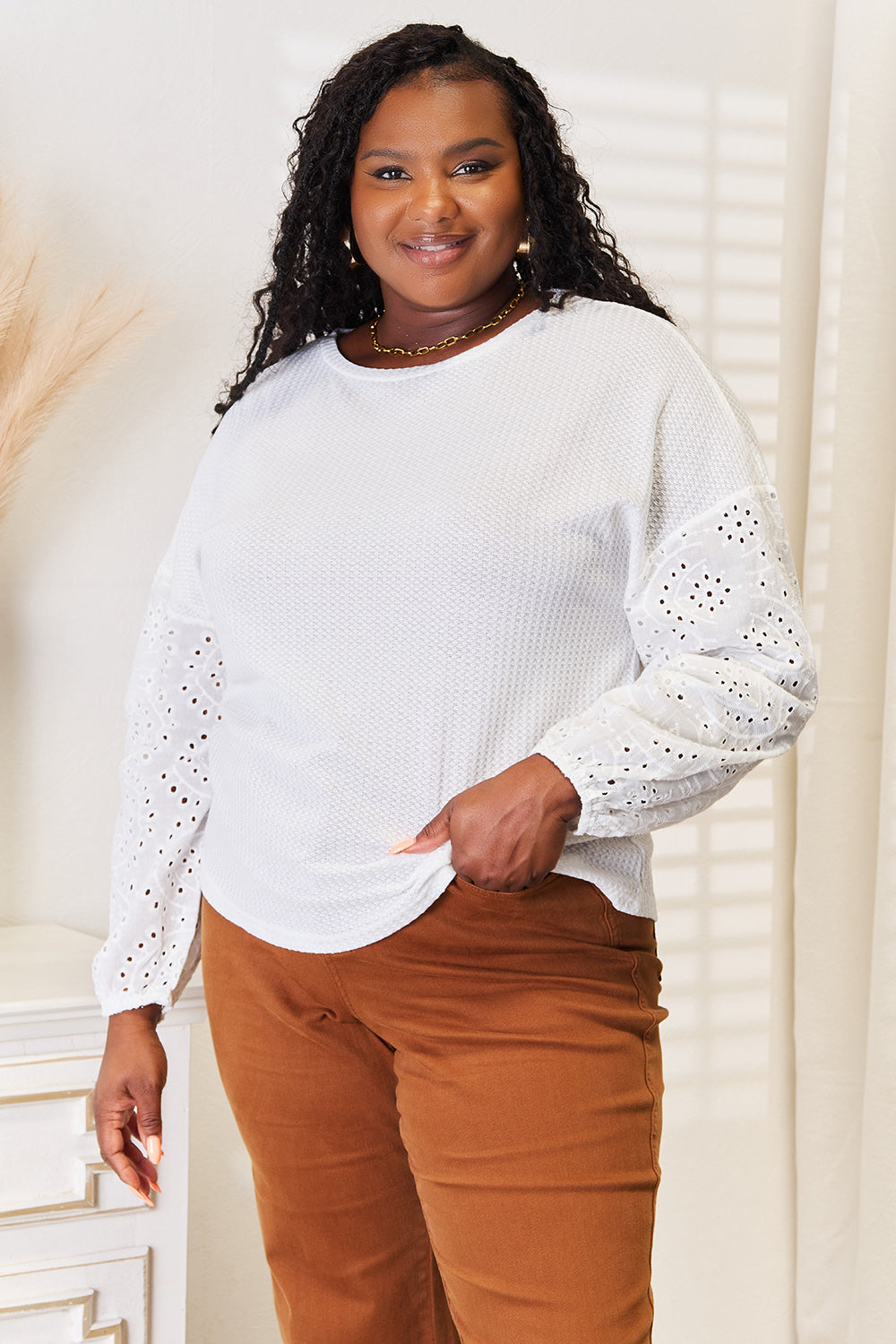 A woman wearing a Shiny Eyelet Dropped Shoulder Round Neck Blouse and brown pants stands in a room with sunlight streaming through blinds. She is smiling and has her left hand on her hip.